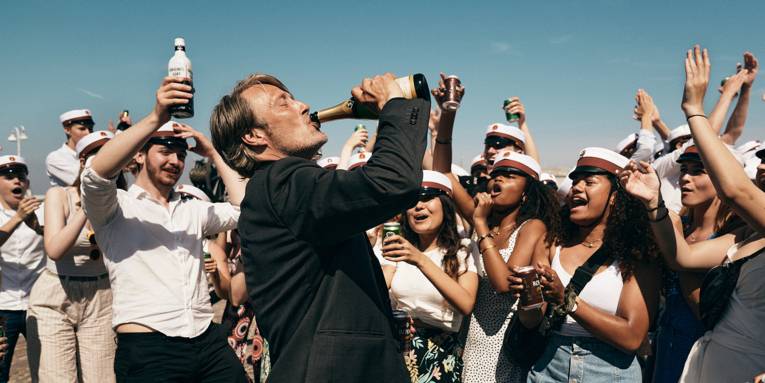 Ein Mann trinkt vor einer ausgelassen feiernden Gruppe Menschen aus einer Champagnerflasche.